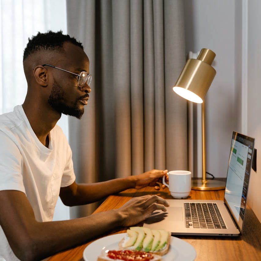 A person is seated at a small desk facing a wall in a hotel-style room, with a hot drink and snack at hand. Image by Mart Production on Pexels.