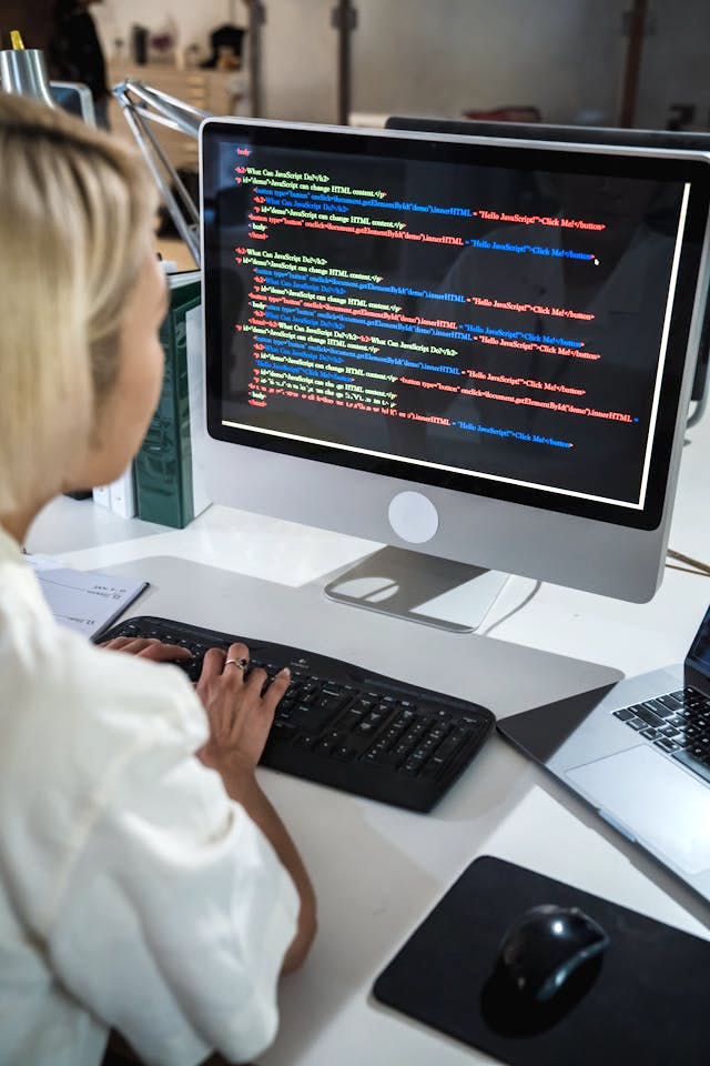 A person is seated at desk behind a monitor showing code. Image by Mizuno Kozuki on Pexels.